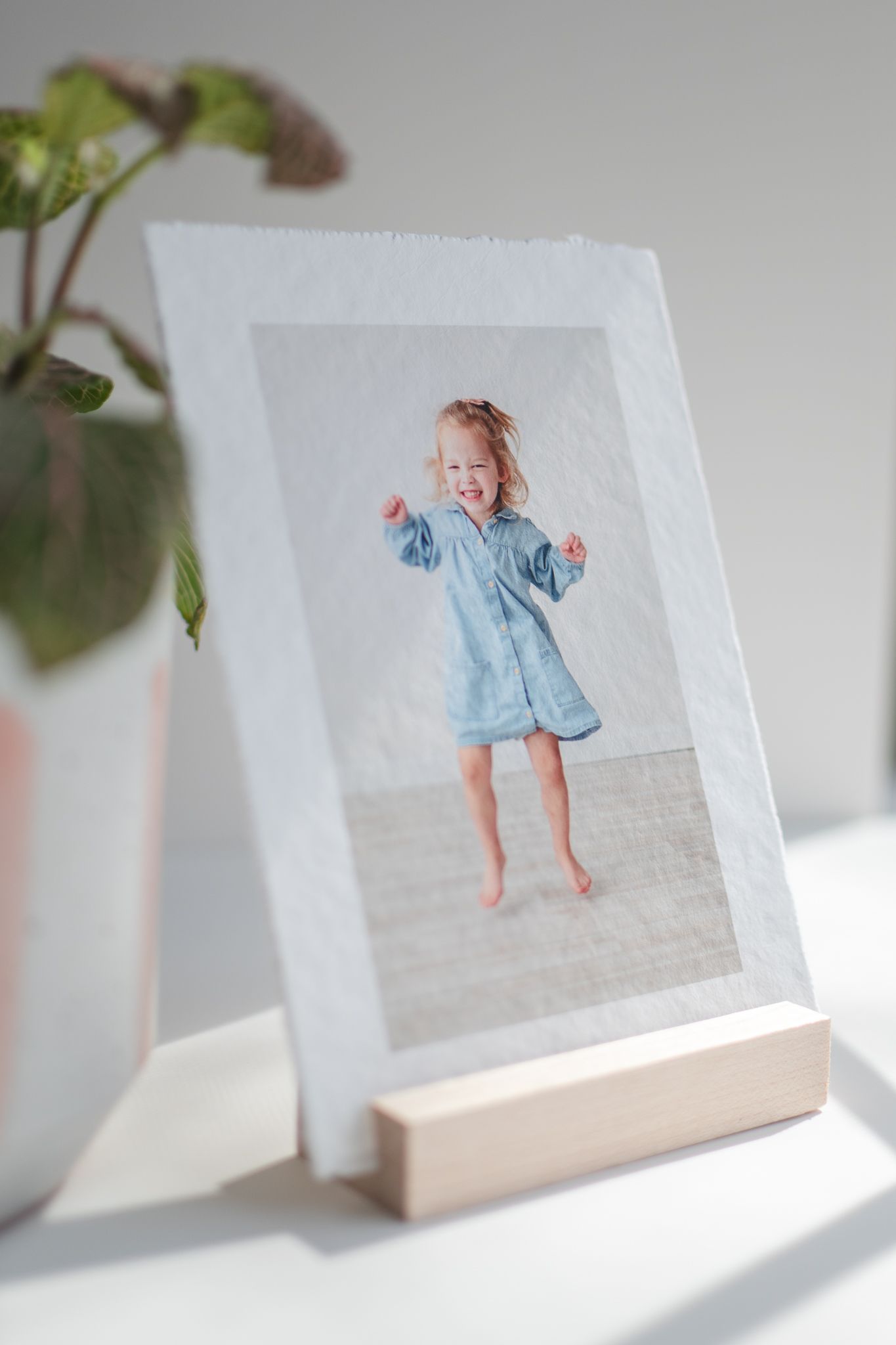 A framed photograph in a simple wooden stand displays a joyful moment in a light-filled setting with a house plant nearby.