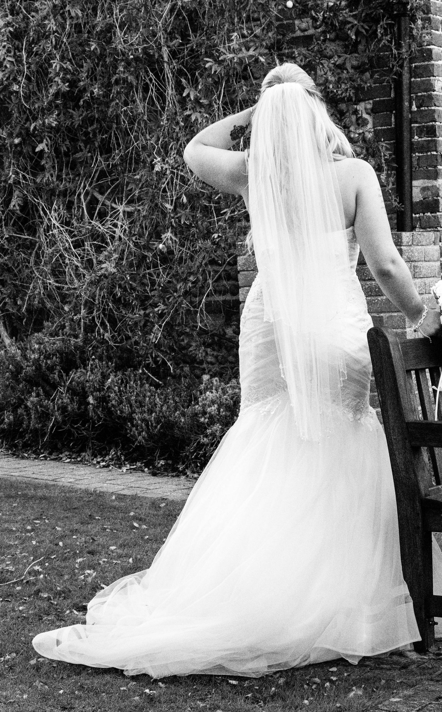 Back view of a bride in a fitted white mermaid wedding dress with long veil standing in a garden setting.