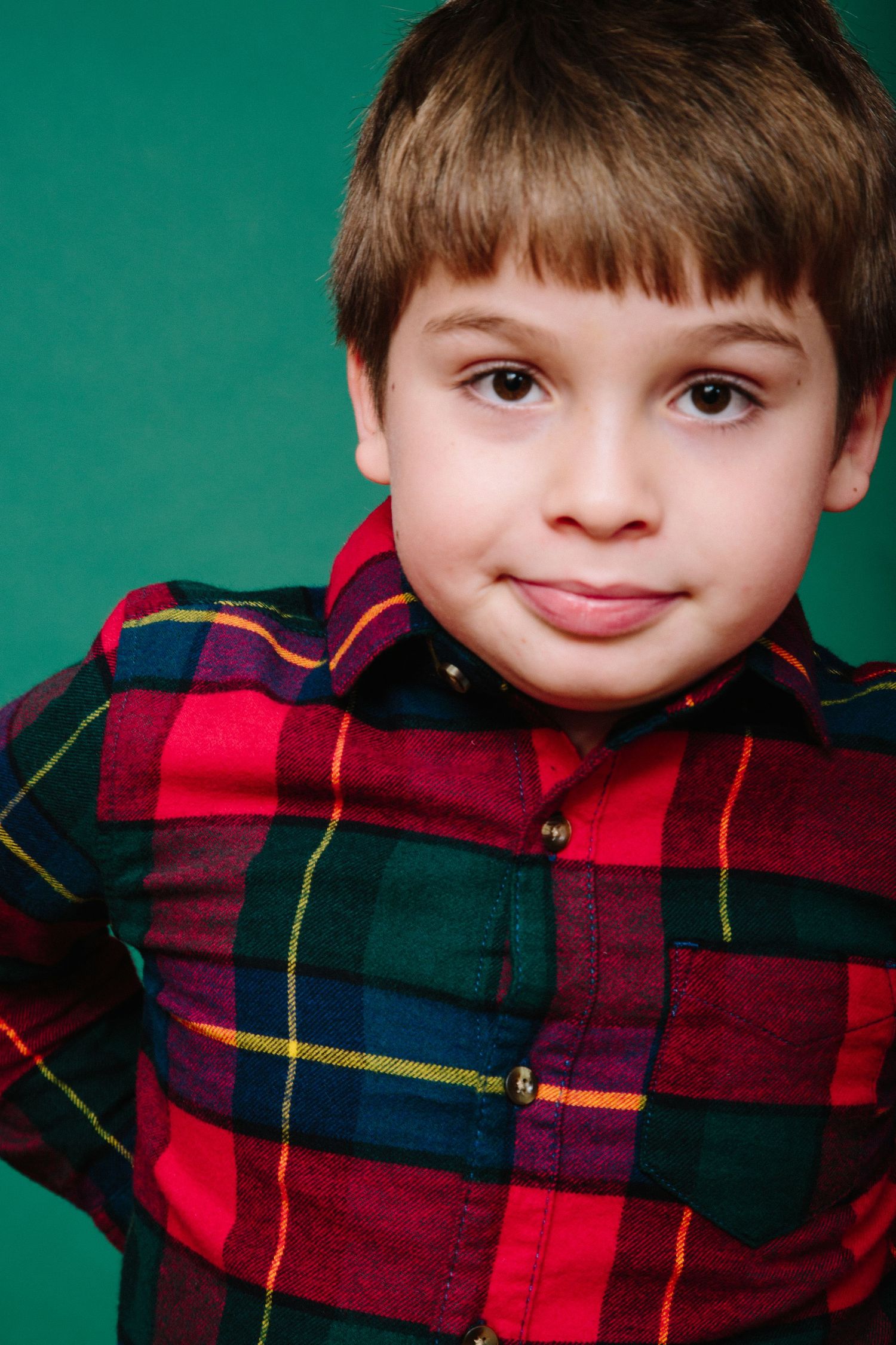 Young child in red plaid shirt smiling at camera against green background.