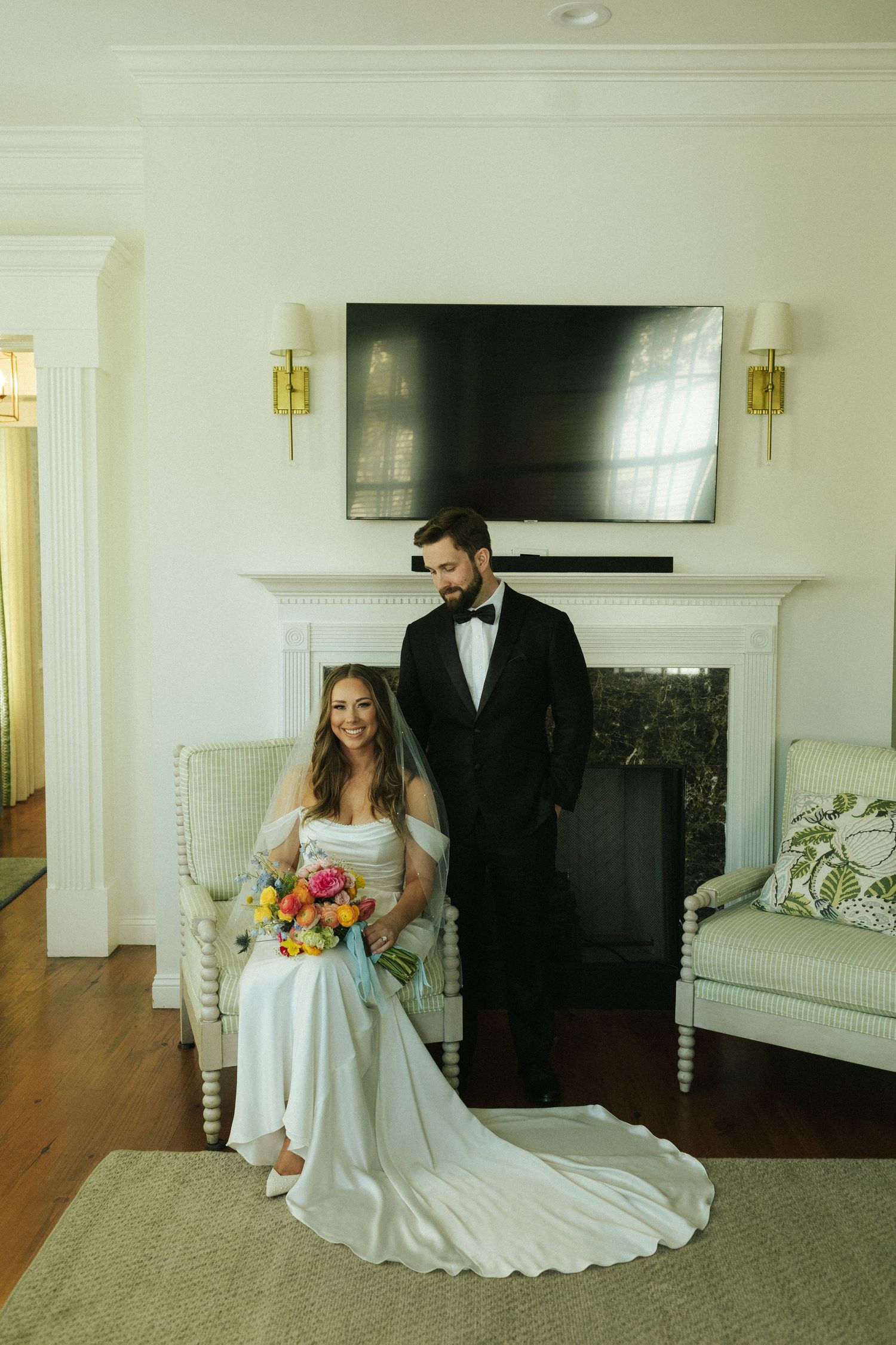 A bride sits in a chair, holding her bouquet in her lap, smiling at the camera while her groom stands behind her, smiling down at her