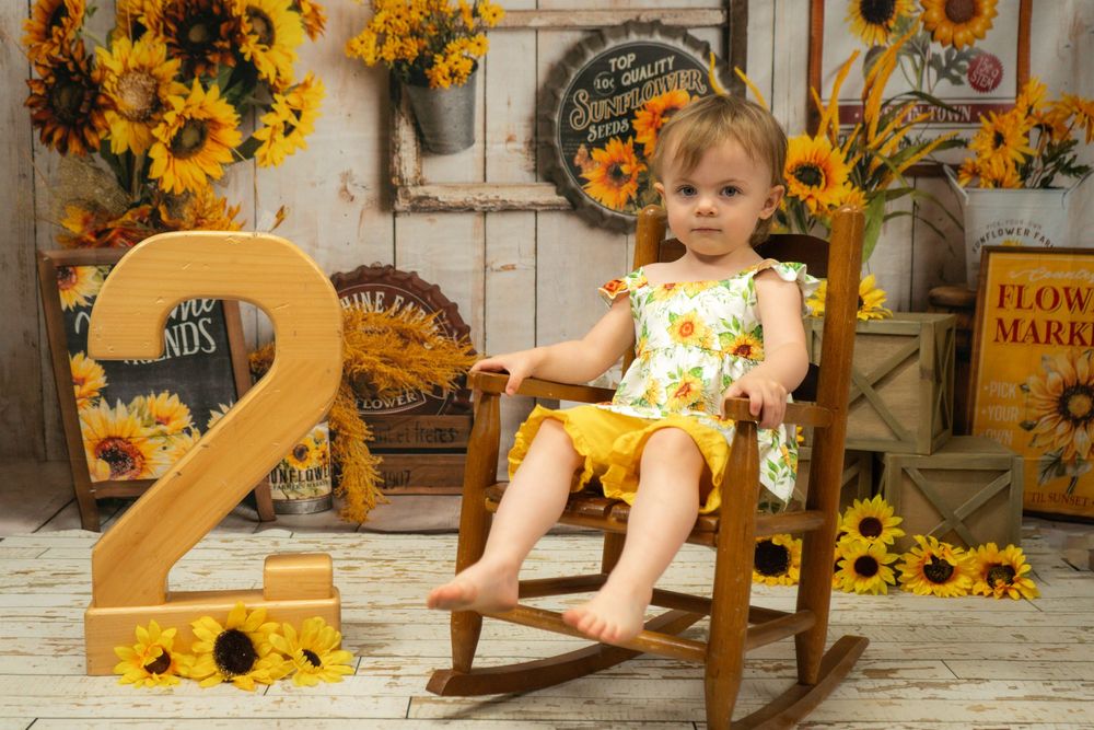 A child sits in a wooden rocking chair surrounded by sunflowers and a large golden number two decoration.