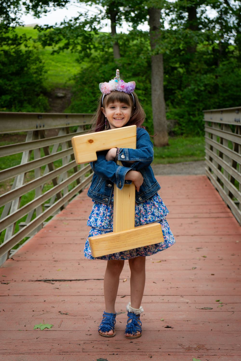 Young child in blue floral dress and denim jacket holds large wooden cross while standing on wooden bridge in park.