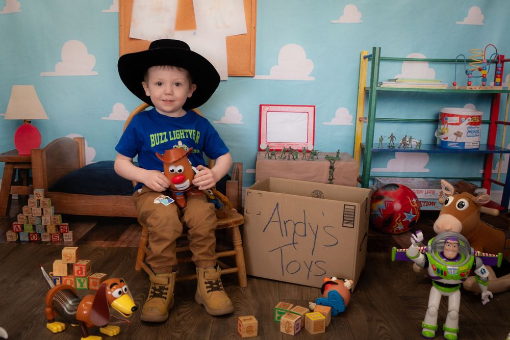 A toddler in cowboy attire sits in a Toy Story themed room with Woody, Buzz Lightyear and other toys around.
