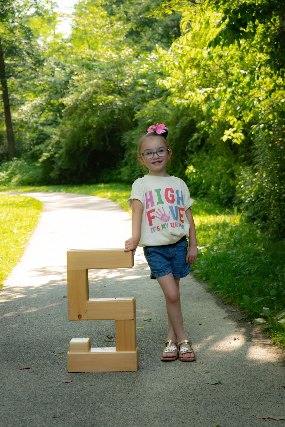 Child standing on a path in a wooded area next to a large decorative wooden number 5 on a sunny day.
