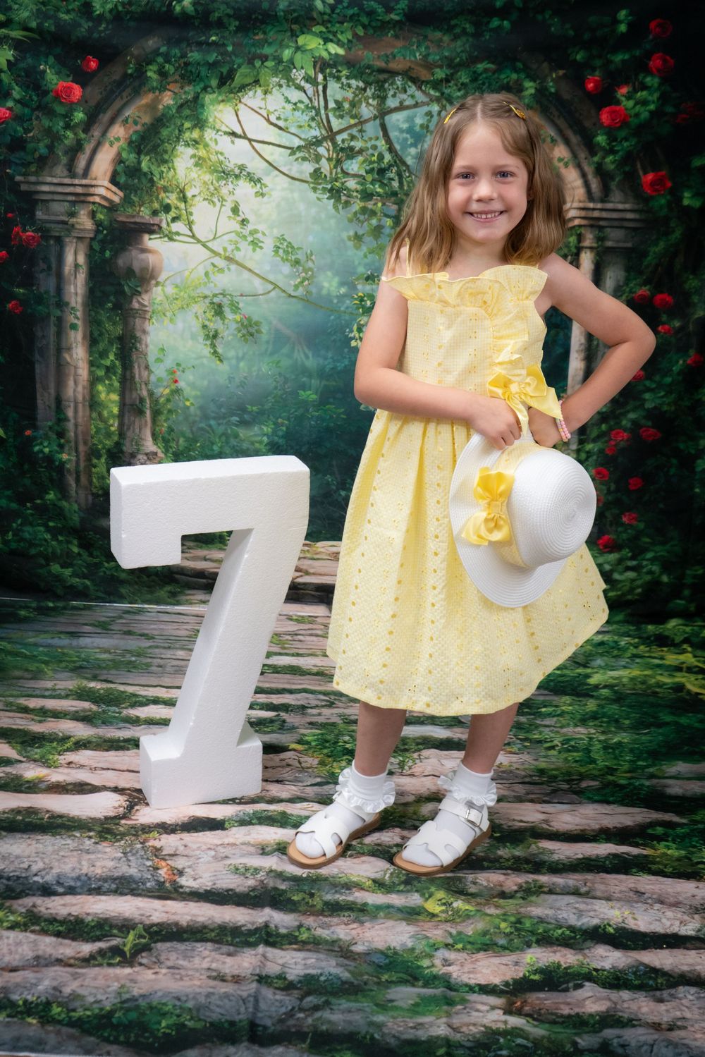 A child in a yellow polka dot dress poses with white 3D number props against a lush floral garden background.