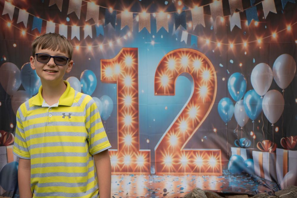 A child in yellow striped shirt and sunglasses poses in front of a light-up number 12 with blue balloons and party decorations.