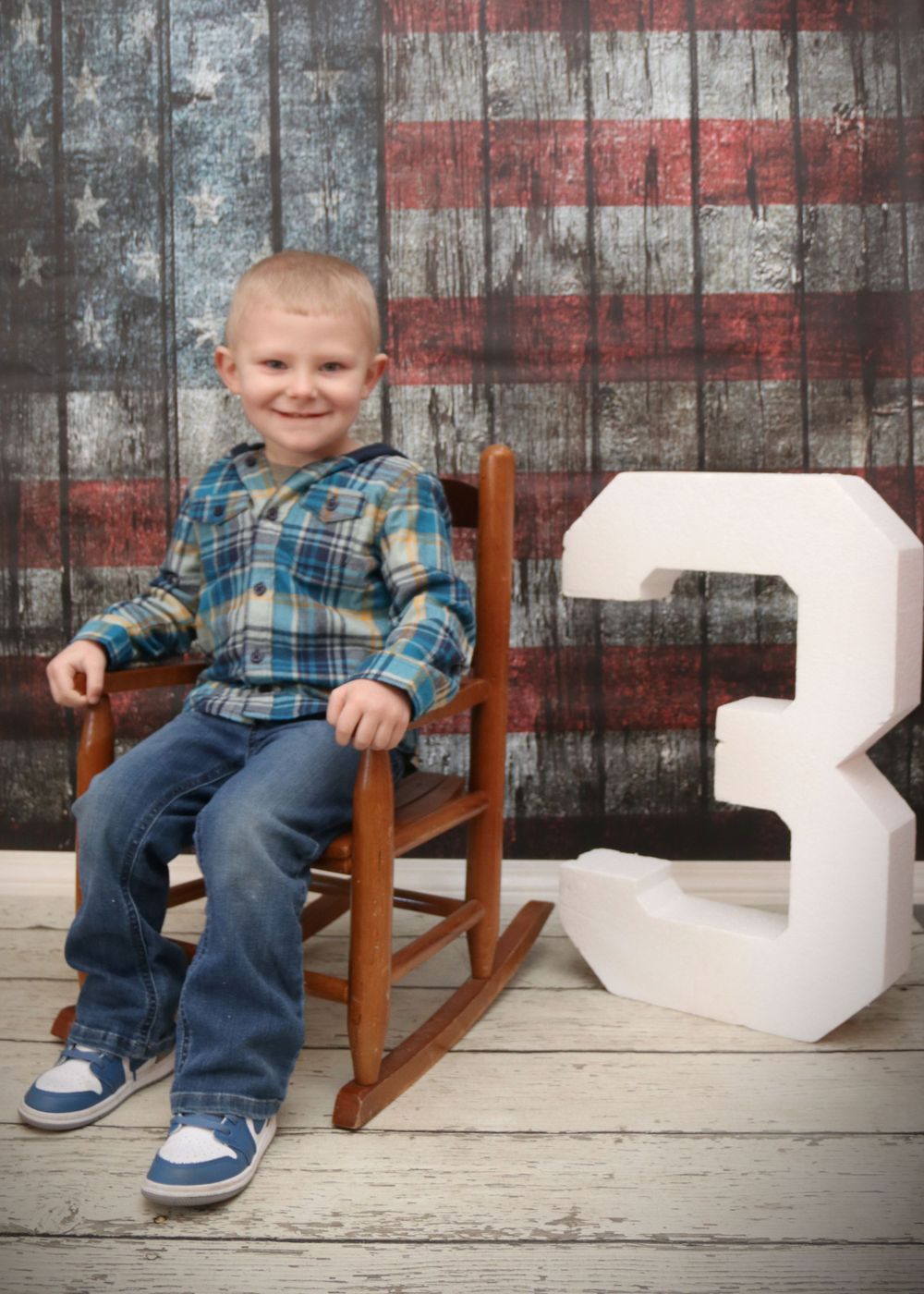 Child in plaid shirt sits in wooden rocking chair beside number 3 against American flag backdrop.
