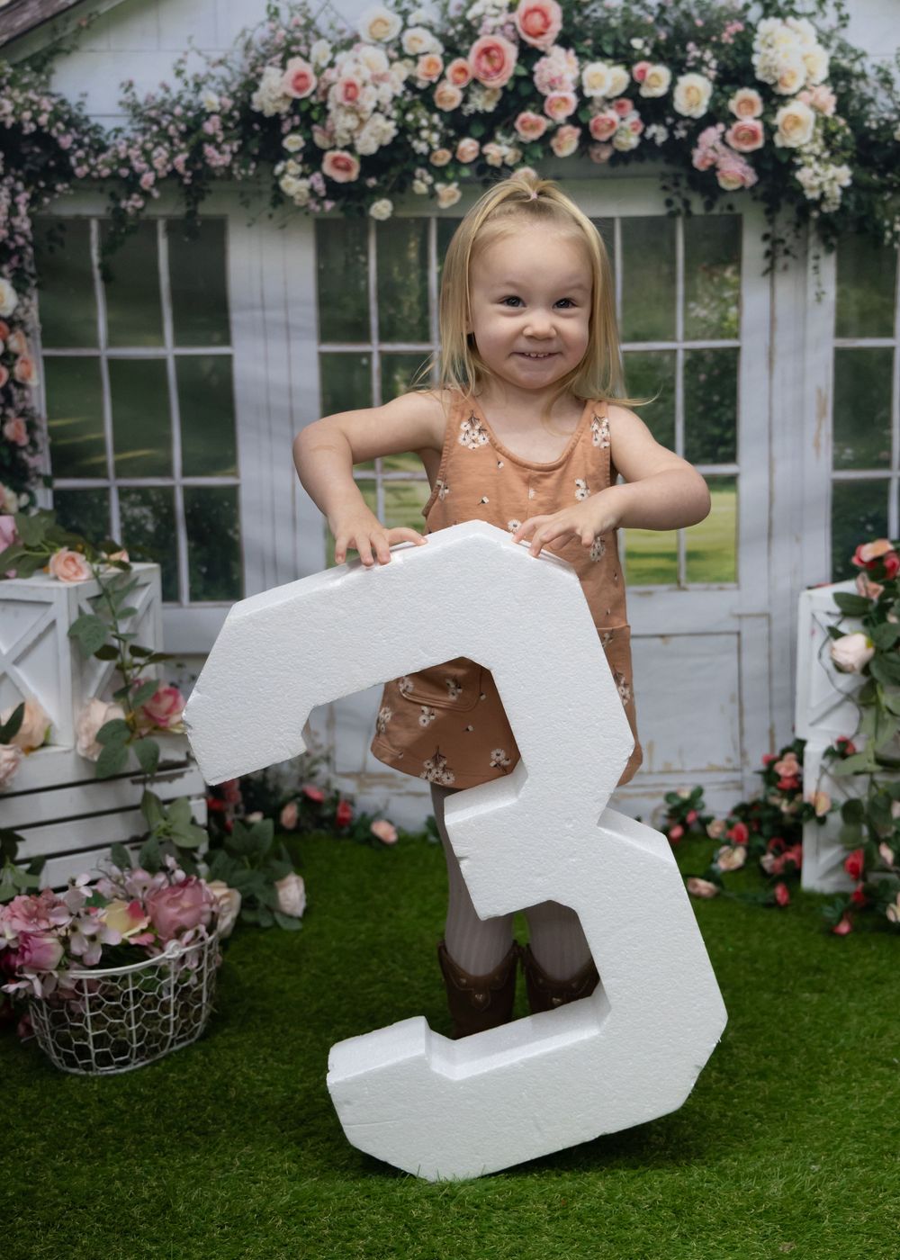 Small child poses with large white number 3 prop surrounded by pink and white roses on floral backdrop display.