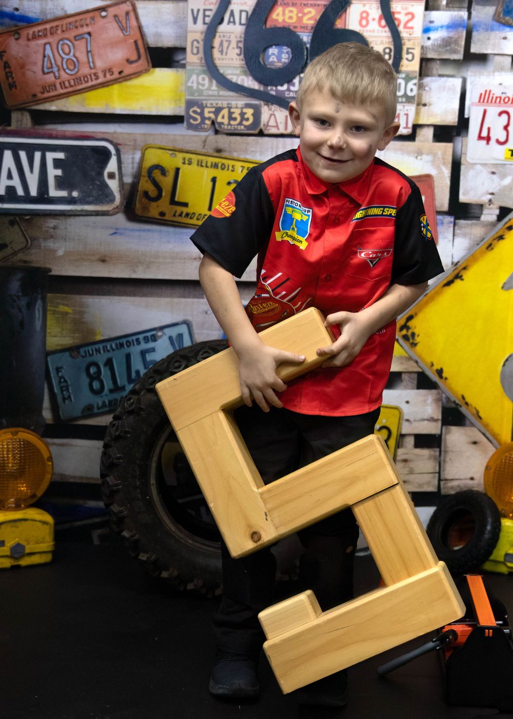 Young person in red vest stands in front of wall of vintage license plates while holding a wooden step stool.