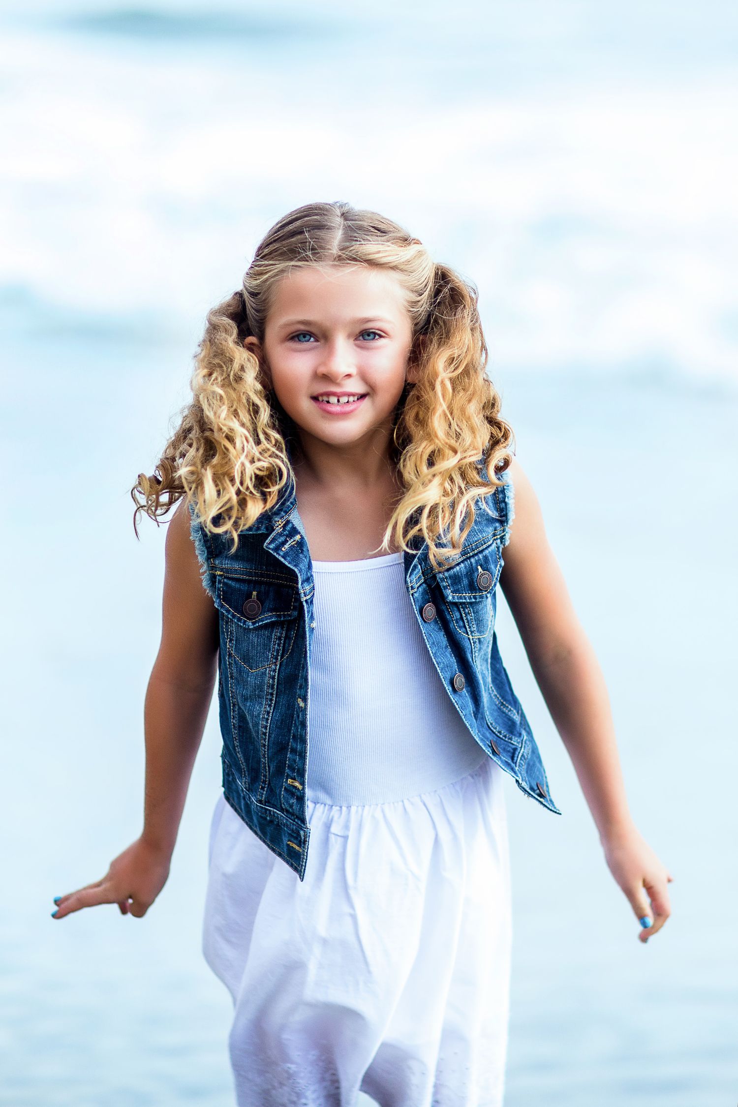 A young child in a white dress and denim vest poses outdoors against a bright blue sky background.
