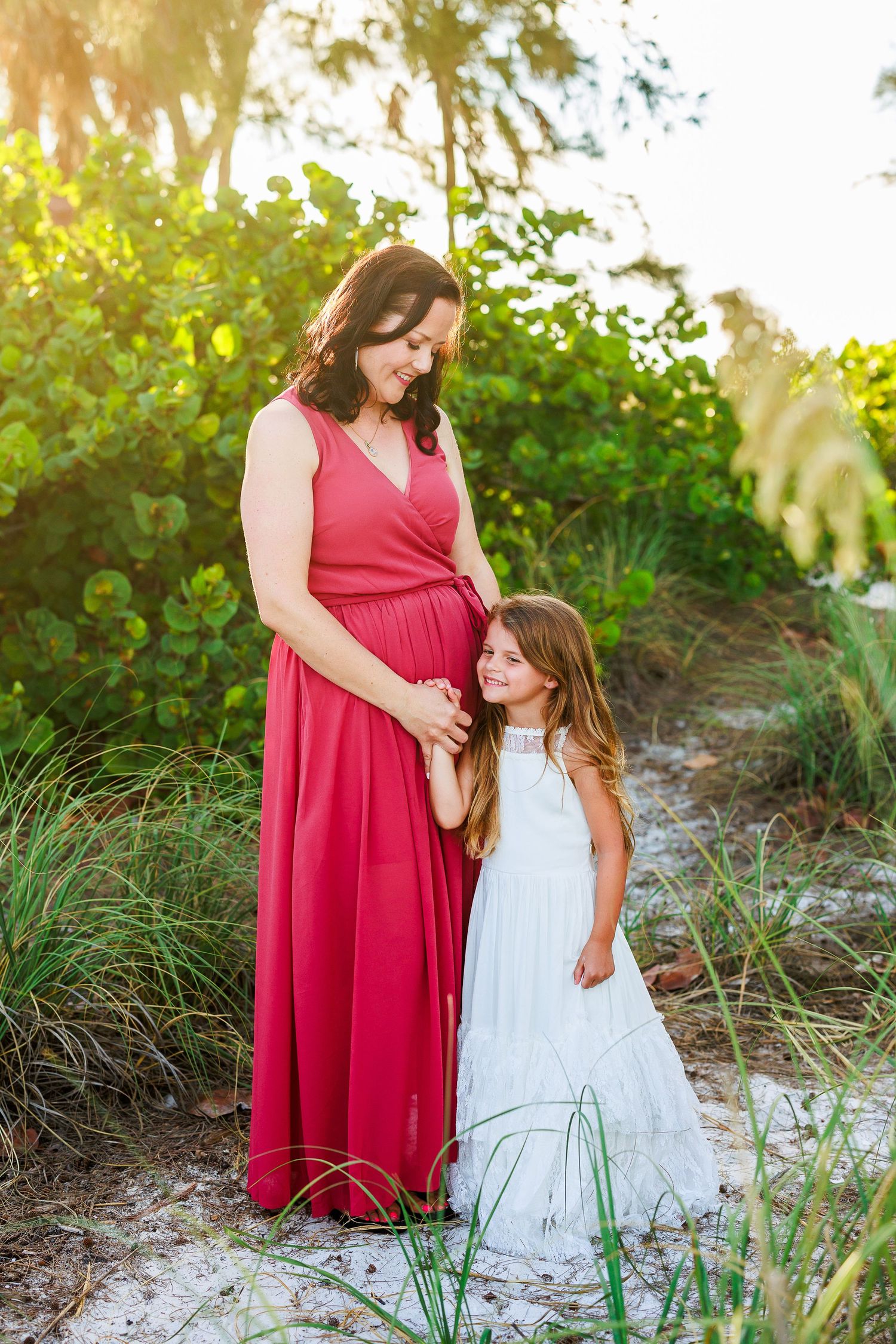 A person in a flowing red dress stands with a young girl in a white dress among beach grass at sunset.