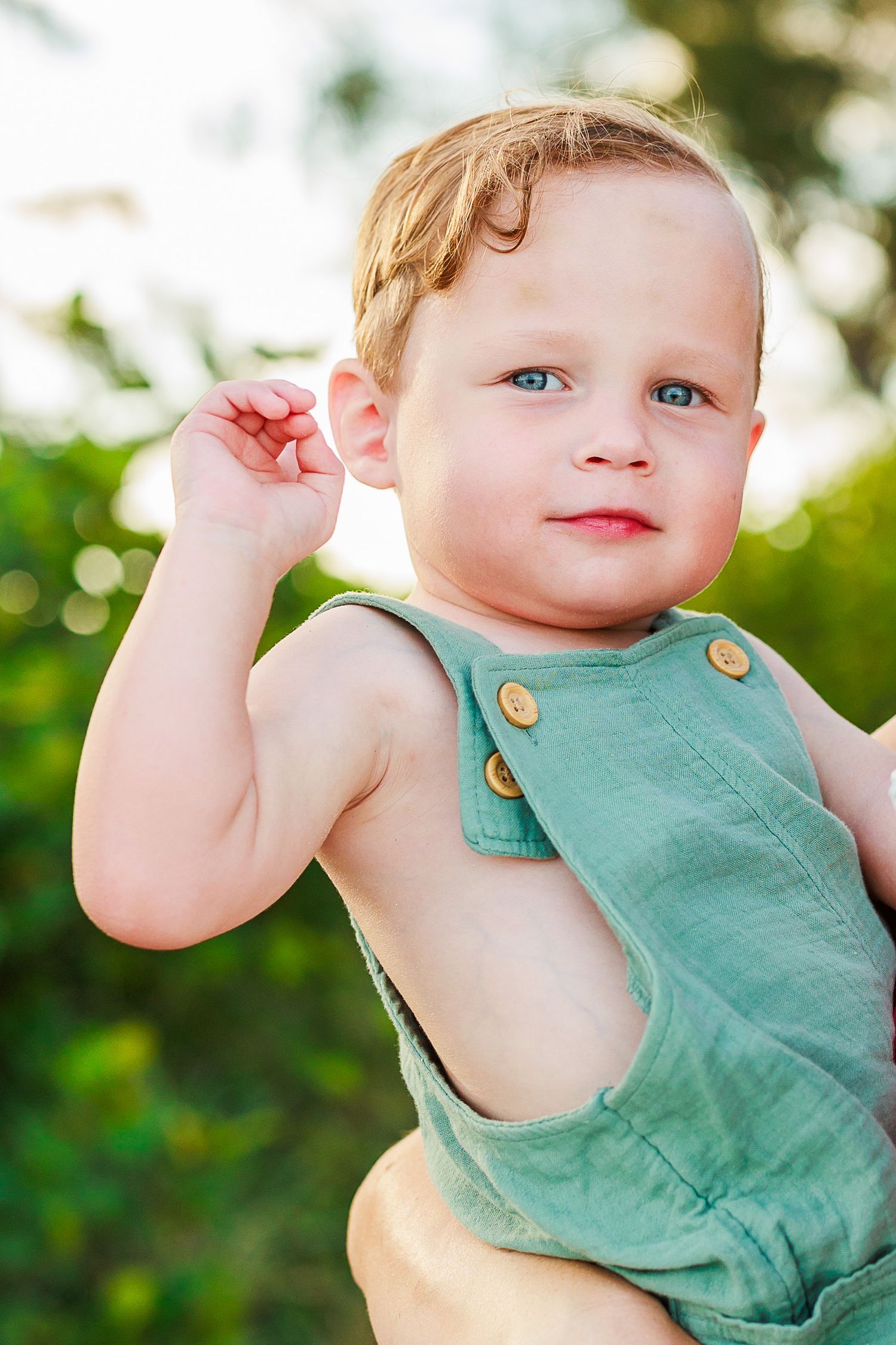 A toddler wearing a mint green romper with gold buttons smiles while playing outdoors in natural lighting.
