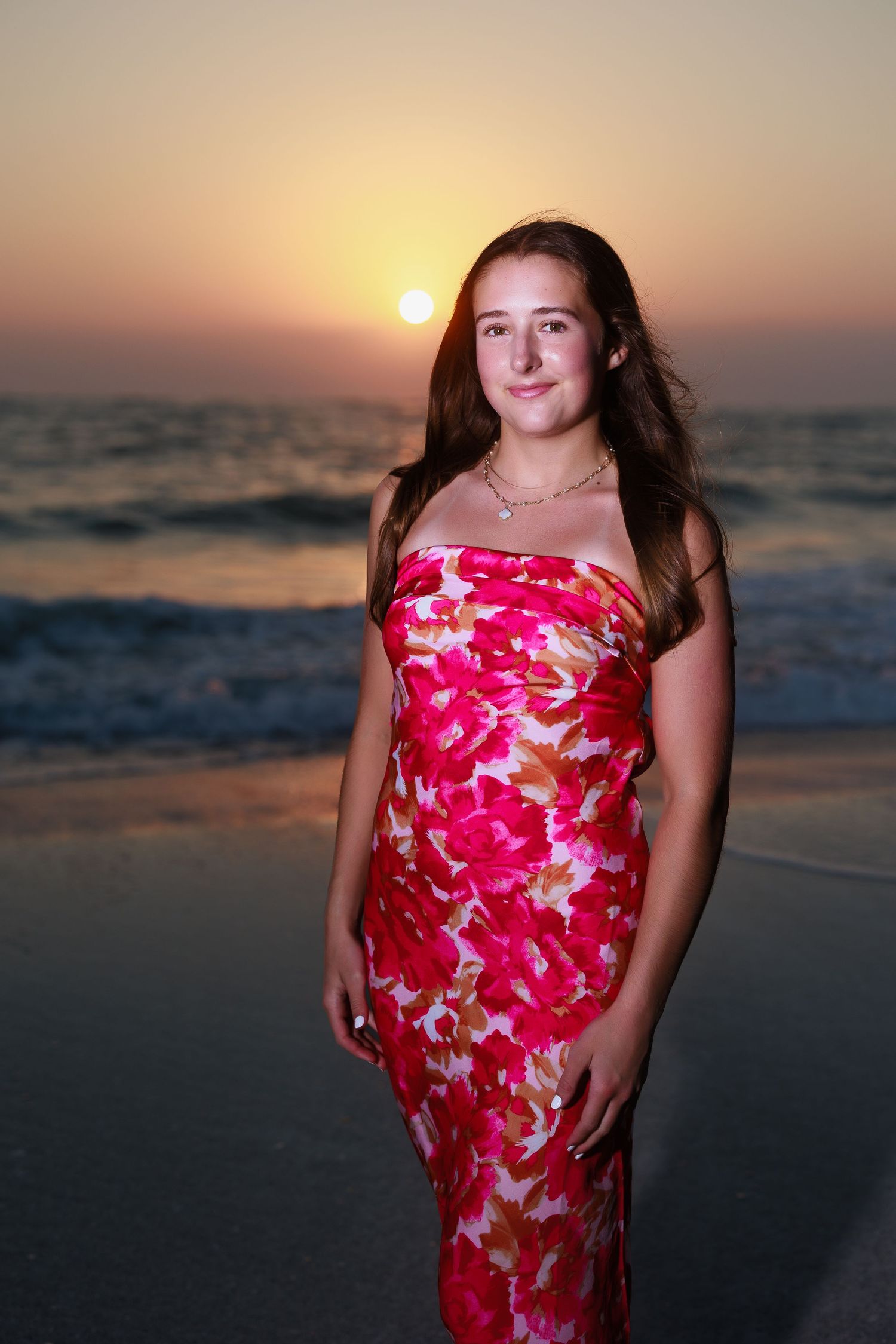 A person in a vibrant red floral dress poses on the beach at sunset with the ocean waves in the background.