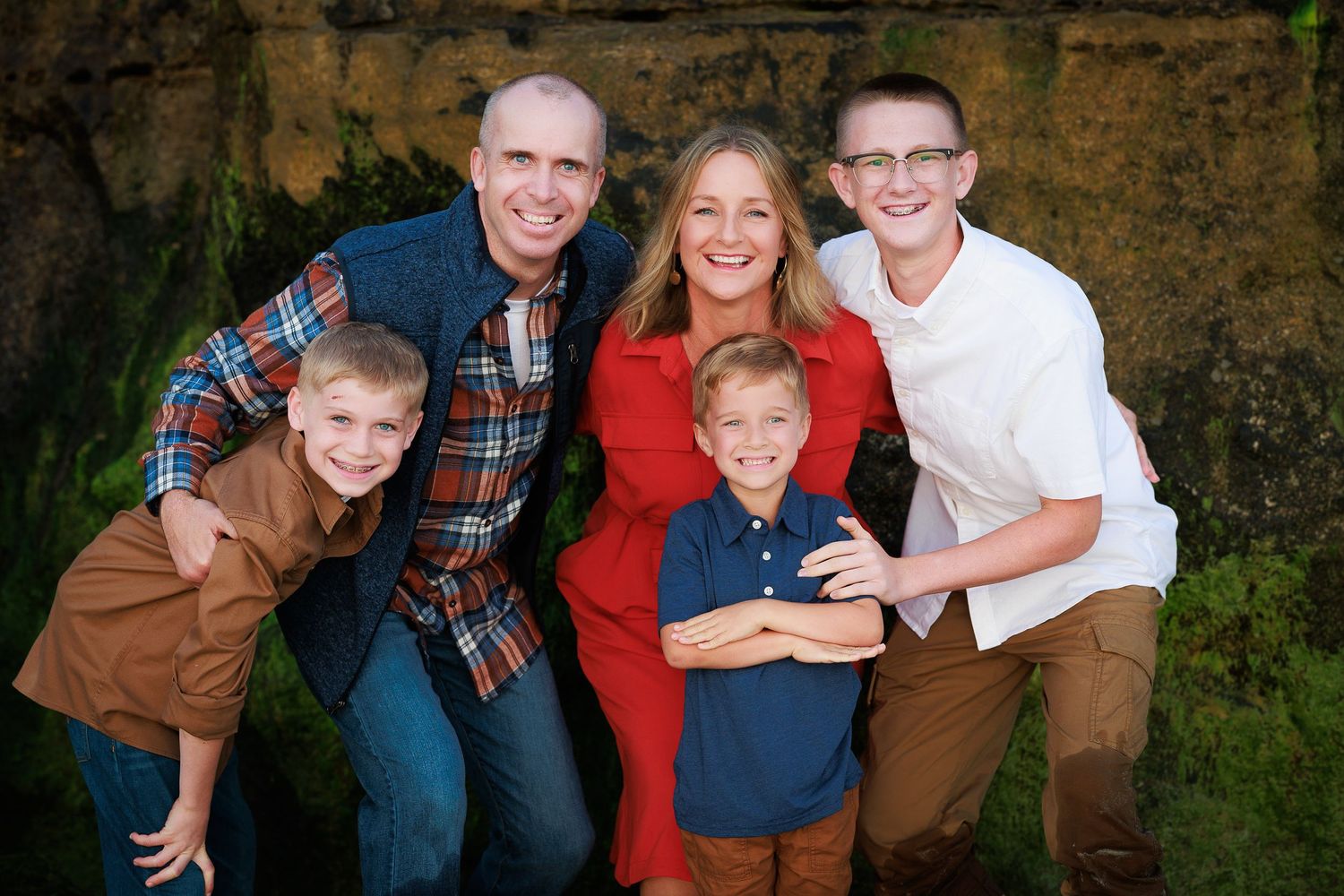 Family portrait with five members wearing coordinated casual clothing posed outdoors against stone wall in natural setting.
