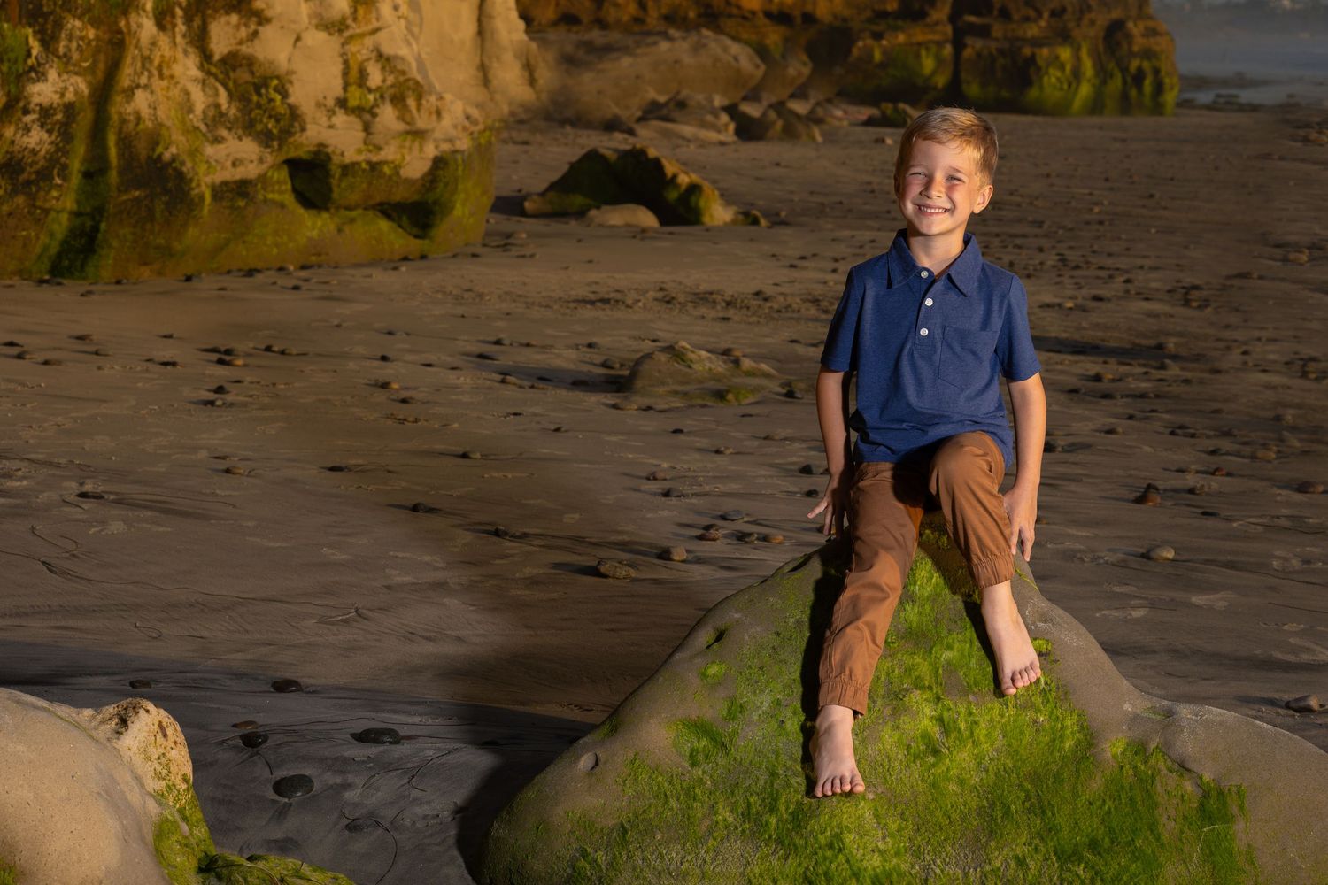 A young individual sits on a moss-covered rock at sunset on a beach with cliffs in the background.