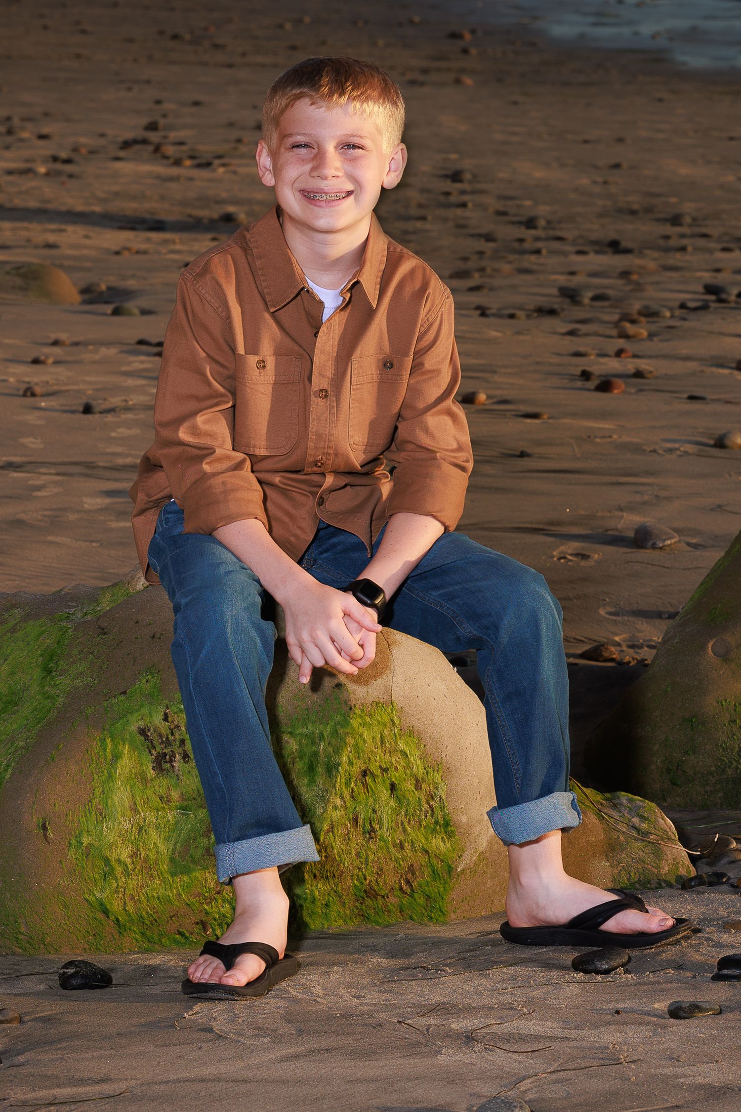 Casual portrait of a youth in a brown shirt and jeans sitting on a mossy rock at the beach wearing flip flops.