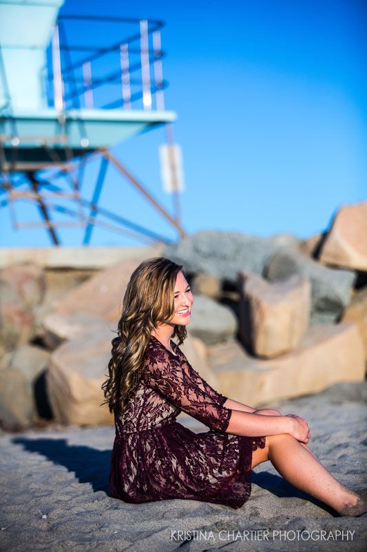 Person in burgundy lace dress sitting on rocks near a lifeguard tower at the beach.