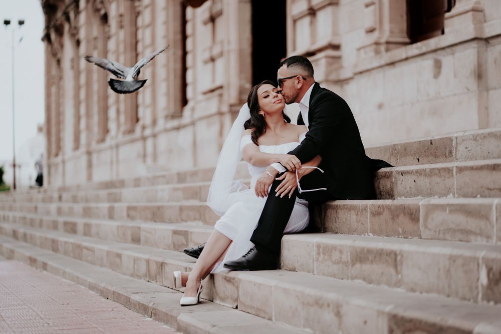 A couple sits on stone steps while a dove flies past in an elegant architectural setting.