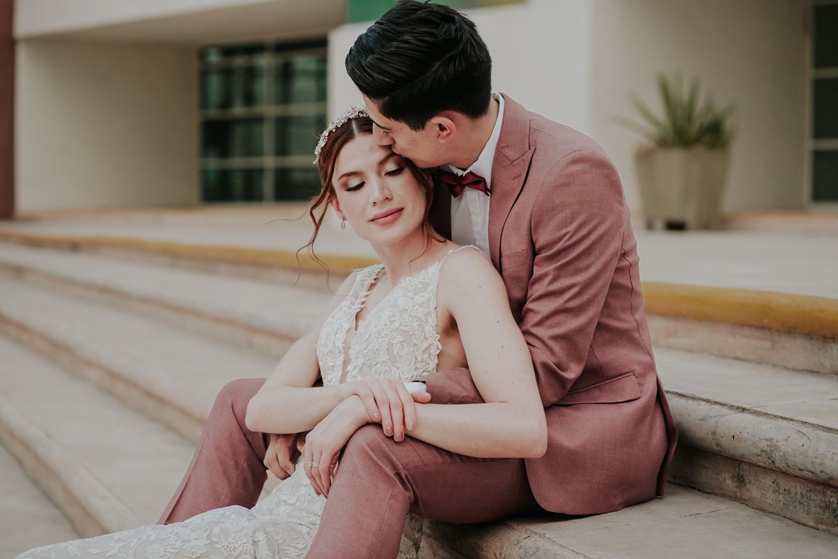 A couple in romantic formal attire sits together on outdoor stone steps sharing intimate moments in a pink-toned photoshoot.