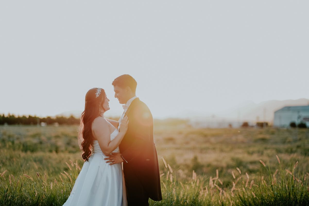 A couple in wedding attire share a romantic moment at sunset in a grassy field with mountains in the background.
