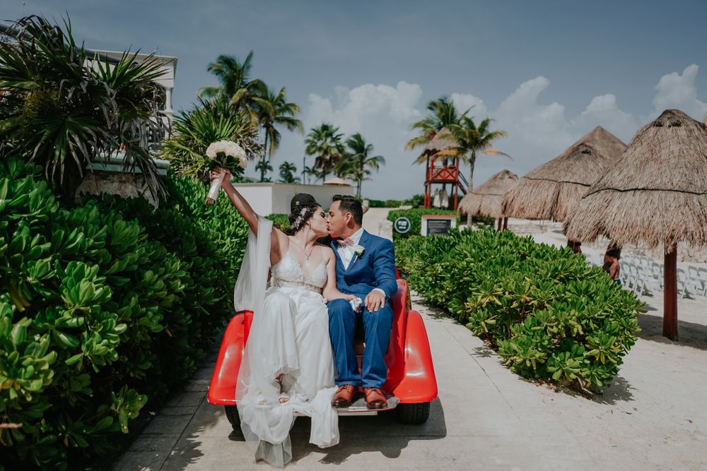 Newlyweds pose on a red golf cart along a tropical path with thatched roof huts.