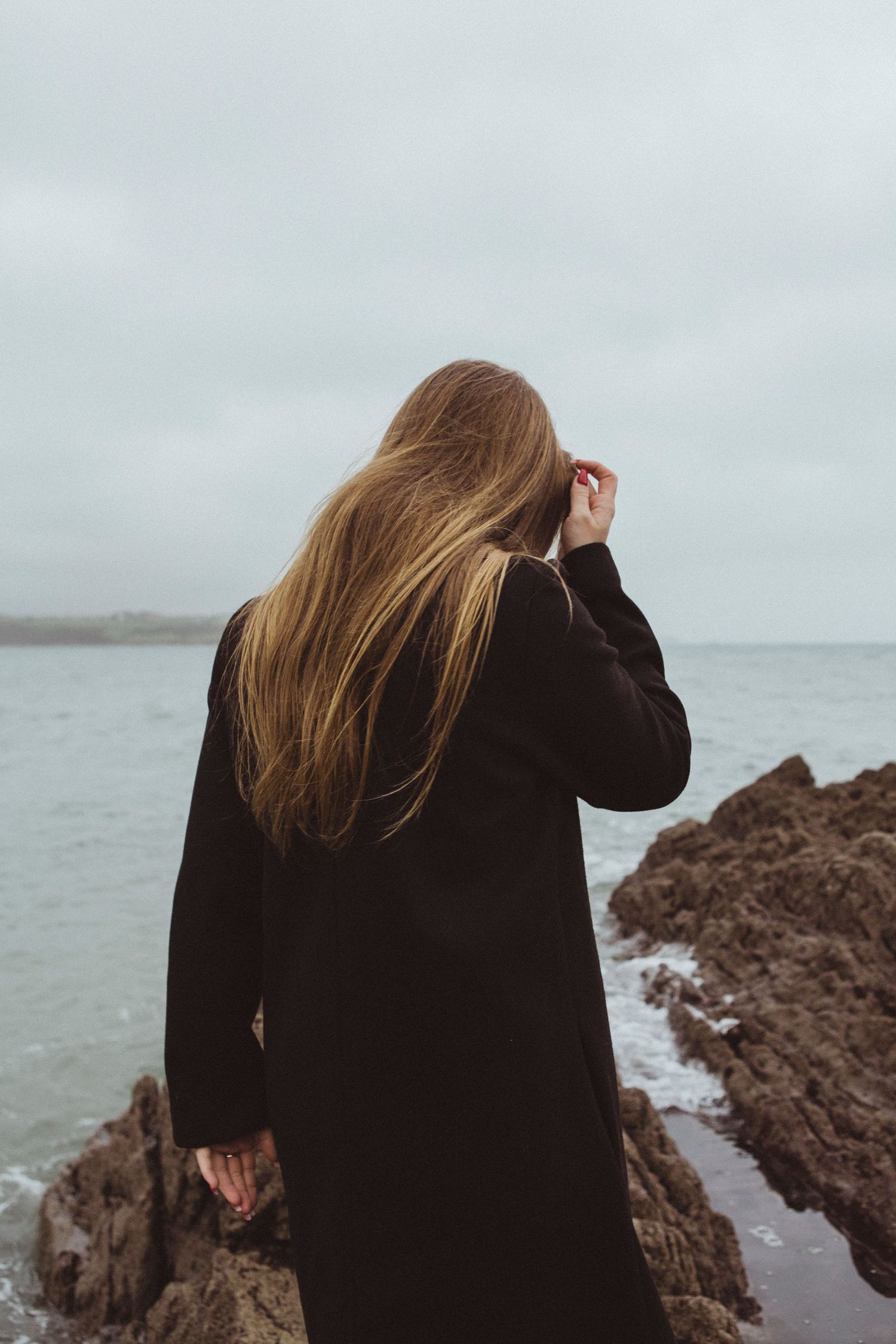 Long-haired figure in black coat stands on rocky coastline with windswept hair against moody grey sky.
