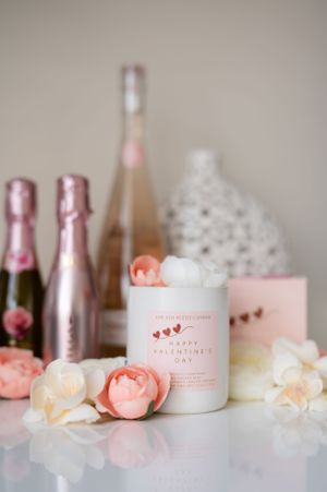 Pink candle with heart design surrounded by roses and champagne bottles in a soft romantic Valentine's Day setting.