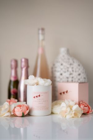 Romantic pink decor arrangement with candle, flowers, and champagne bottles on white surface against neutral background.