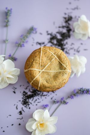 Rustic scone with cross pattern on top arranged with white daffodils and dried tea leaves on purple surface.