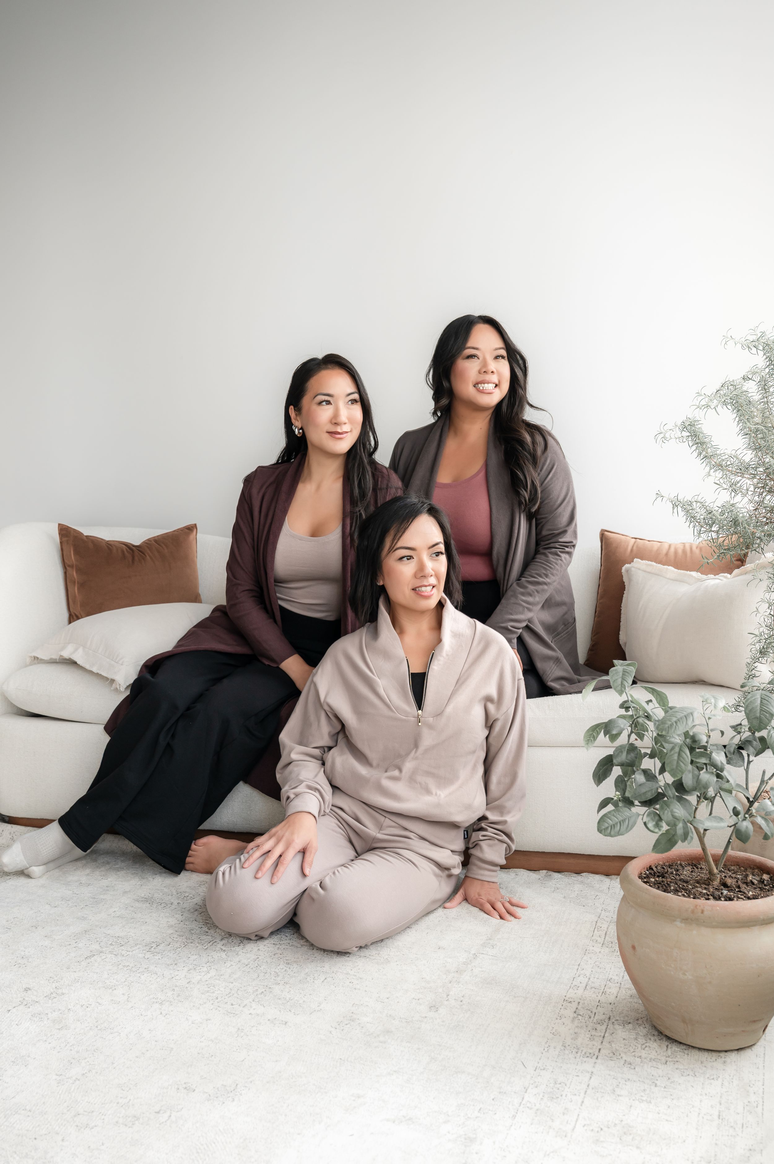 Three women in cozy loungewear relax together on a white couch with decorative plants in the background.