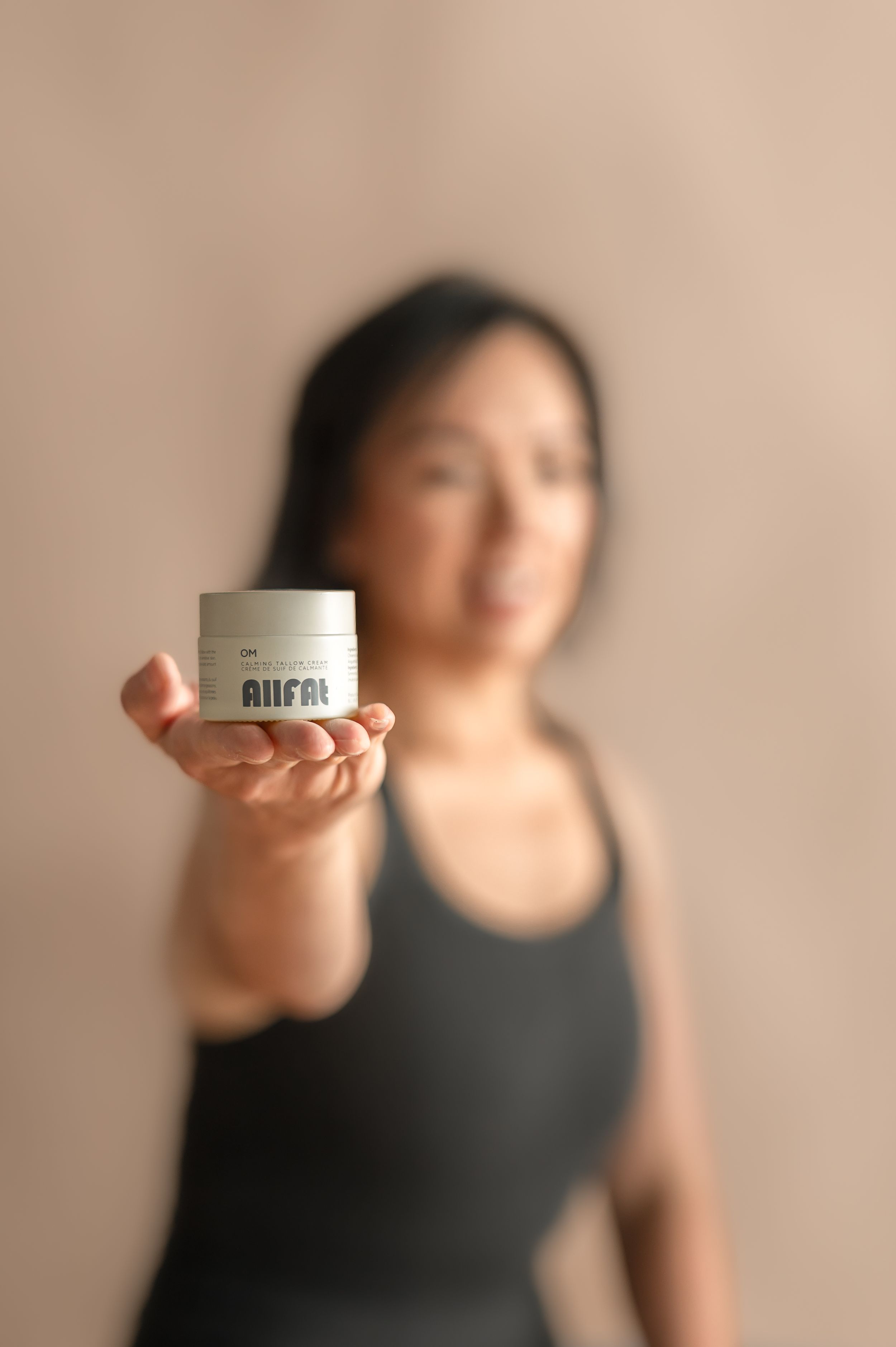 Woman in black tank top holding up a small jar against blurred beige background.
