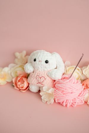 A white plush toy with pink yarn and delicate peach roses arranged on a soft pink background.