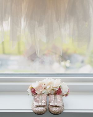 Sparkly rose gold baby shoes decorated with flowers on a white windowsill.