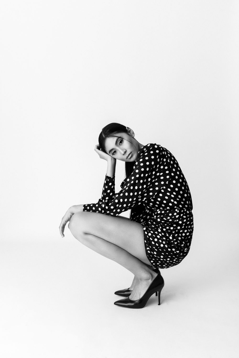 A person in a polka dot dress and heels crouches against a white background in a black and white fashion photo.