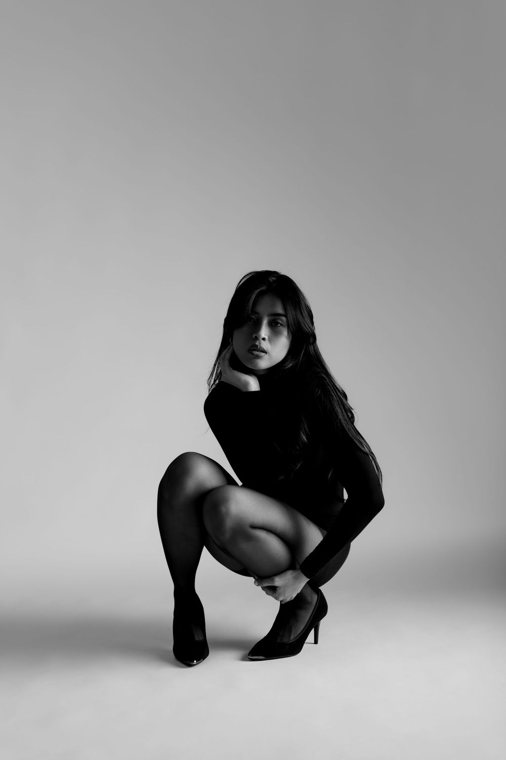 Black and white studio photo of a woman in a black outfit and heels crouching gracefully against a light background.
