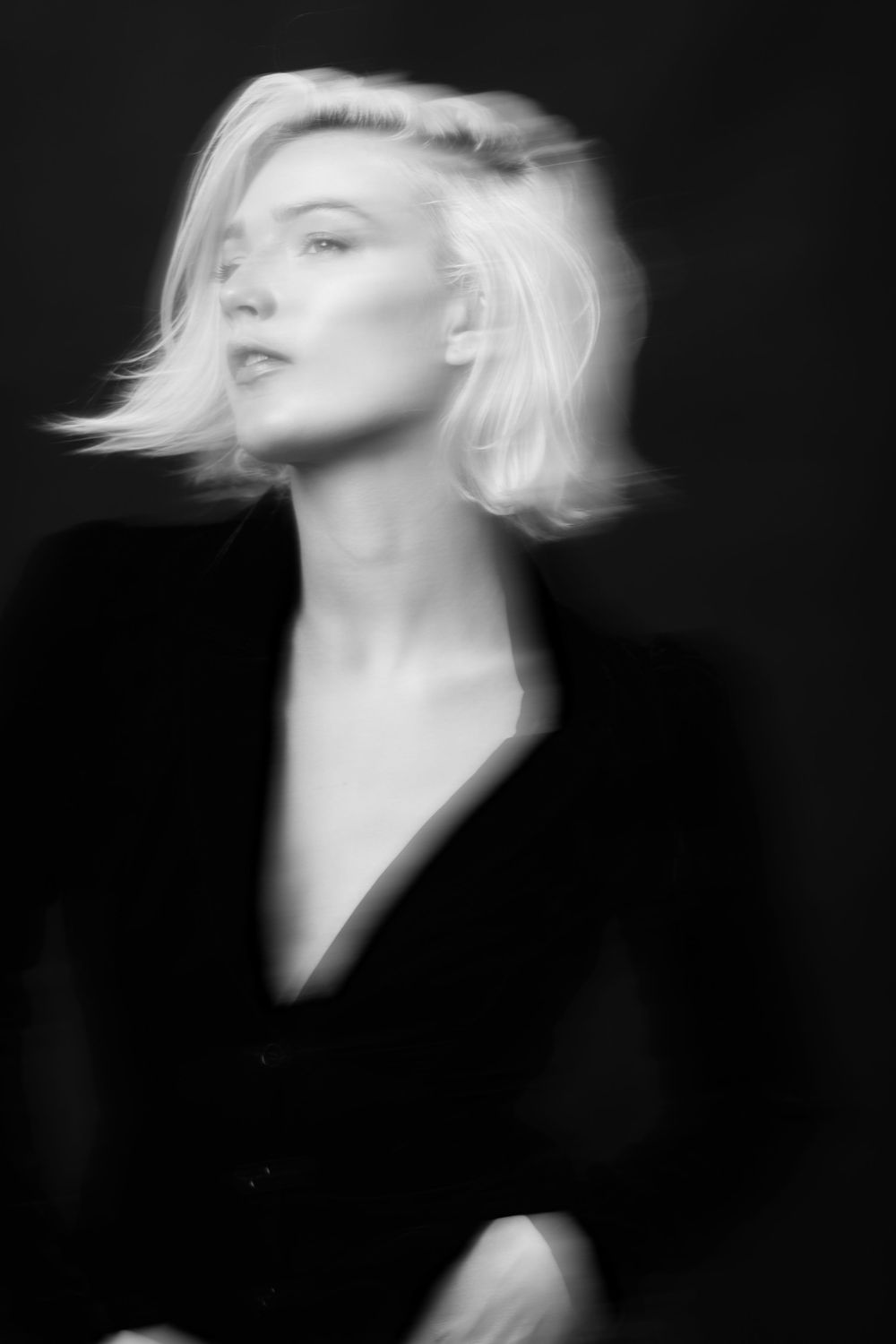 Black and white portrait of a blonde woman in a black blazer with motion blur effect on dark background.