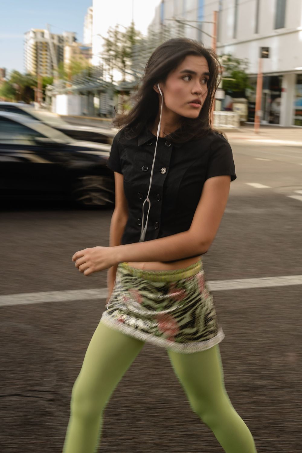 Young woman in black top, green leggings, and mini skirt crosses a busy urban street while listening to earphones.