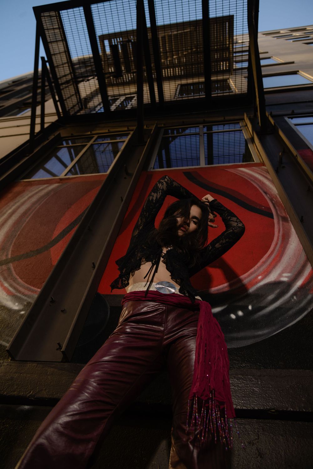 Woman in red leather pants and black lace top posing on urban fire escape.