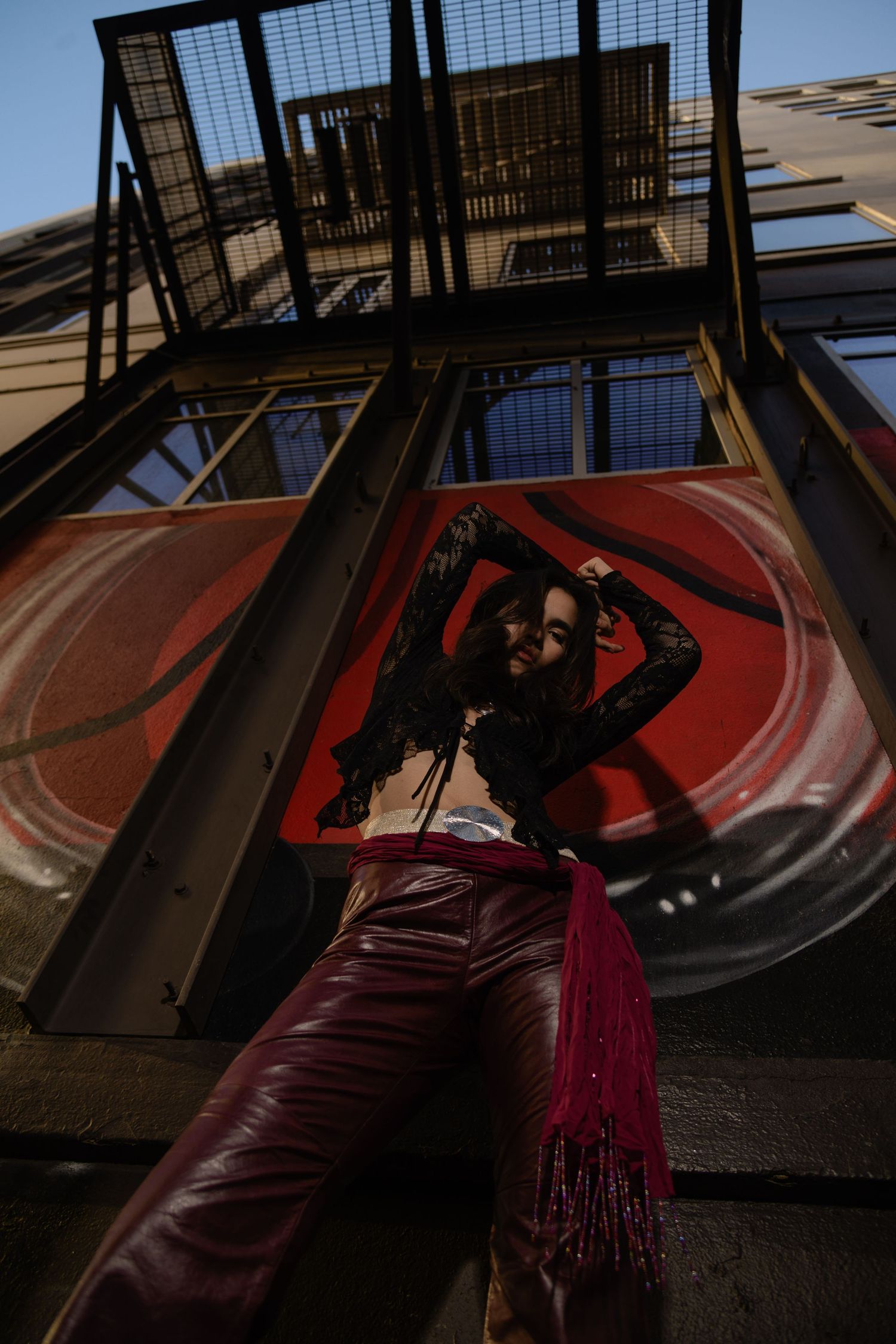 Woman in red leather pants and black lace top posing on urban fire escape.