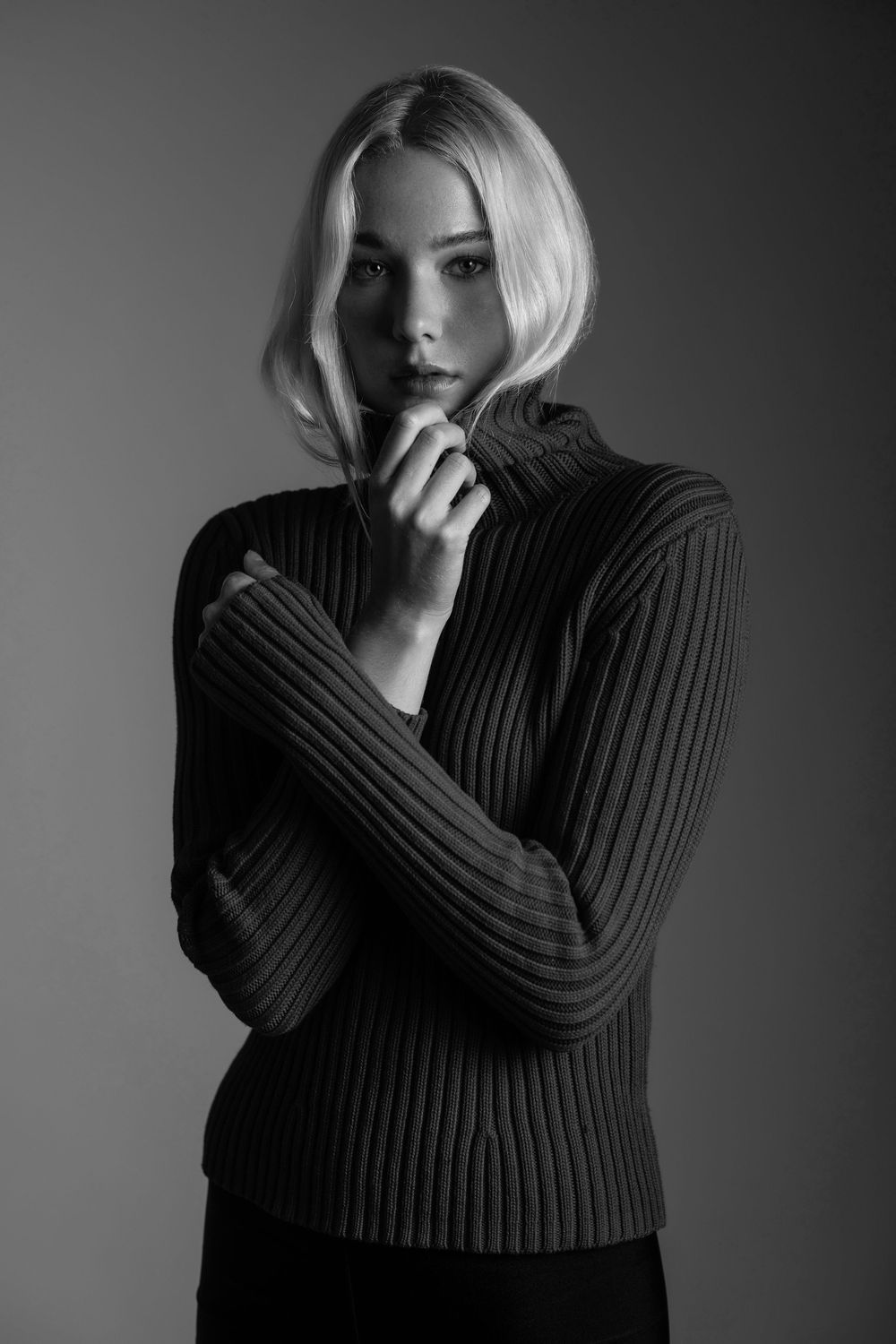 Blonde woman in ribbed turtleneck sweater posing in black and white studio portrait.