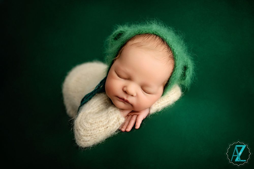Professional newborn photo of sleeping baby wearing green bonnet on dark green background.