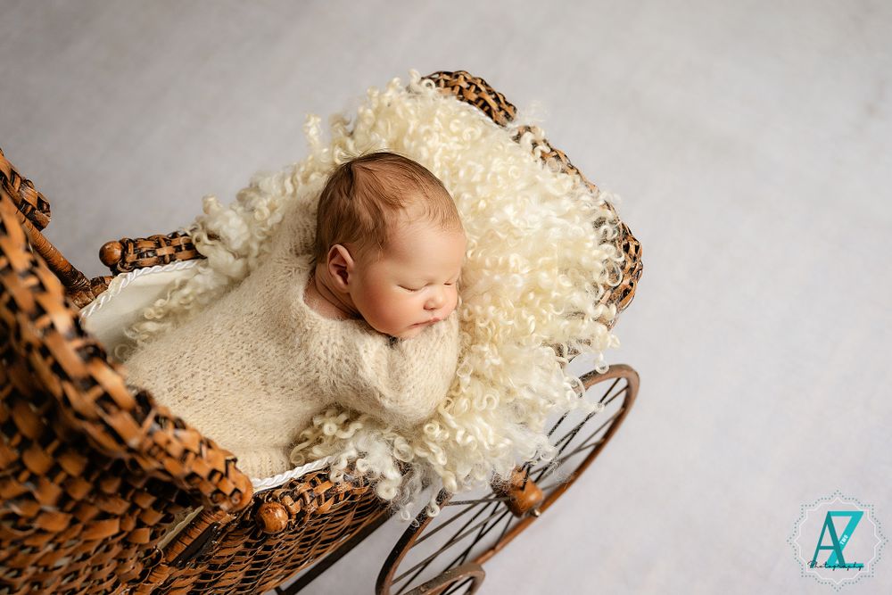 Vintage-style newborn photo with baby nestled in antique wicker carriage with cream blanket.