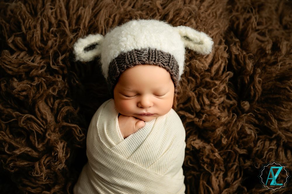 Newborn wearing white knit bear hat swaddled in white wrap on brown textured blanket.