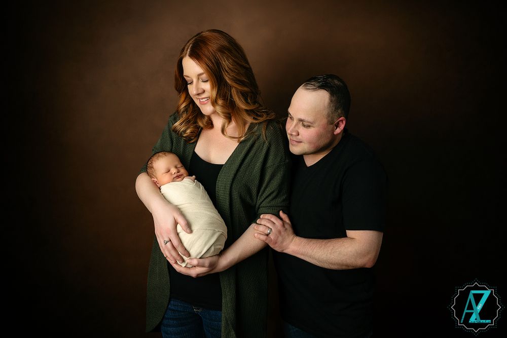 Family portrait against dark background with couple in casual attire holding swaddled infant.