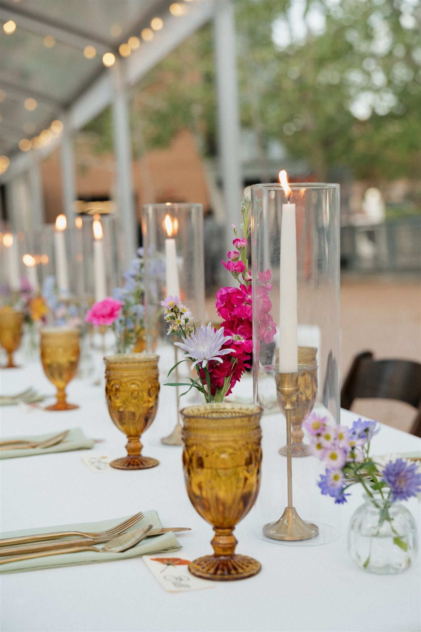 Elegant table setting with amber goblets, tall glass candleholders, and pink floral arrangements on a white tablecloth.