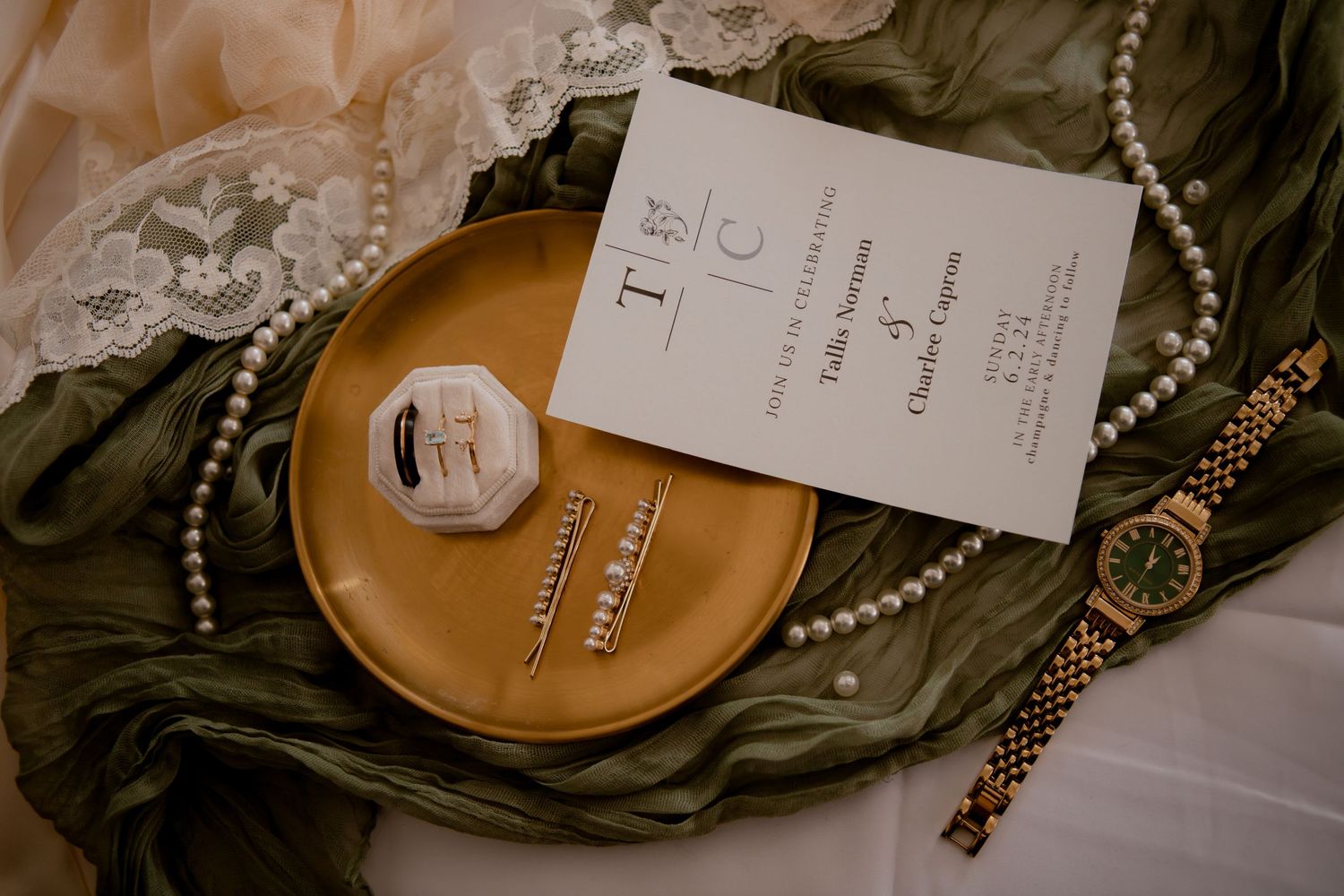 Wedding details flatlay with invitation, rings, and pearls on metallic gold plate.
