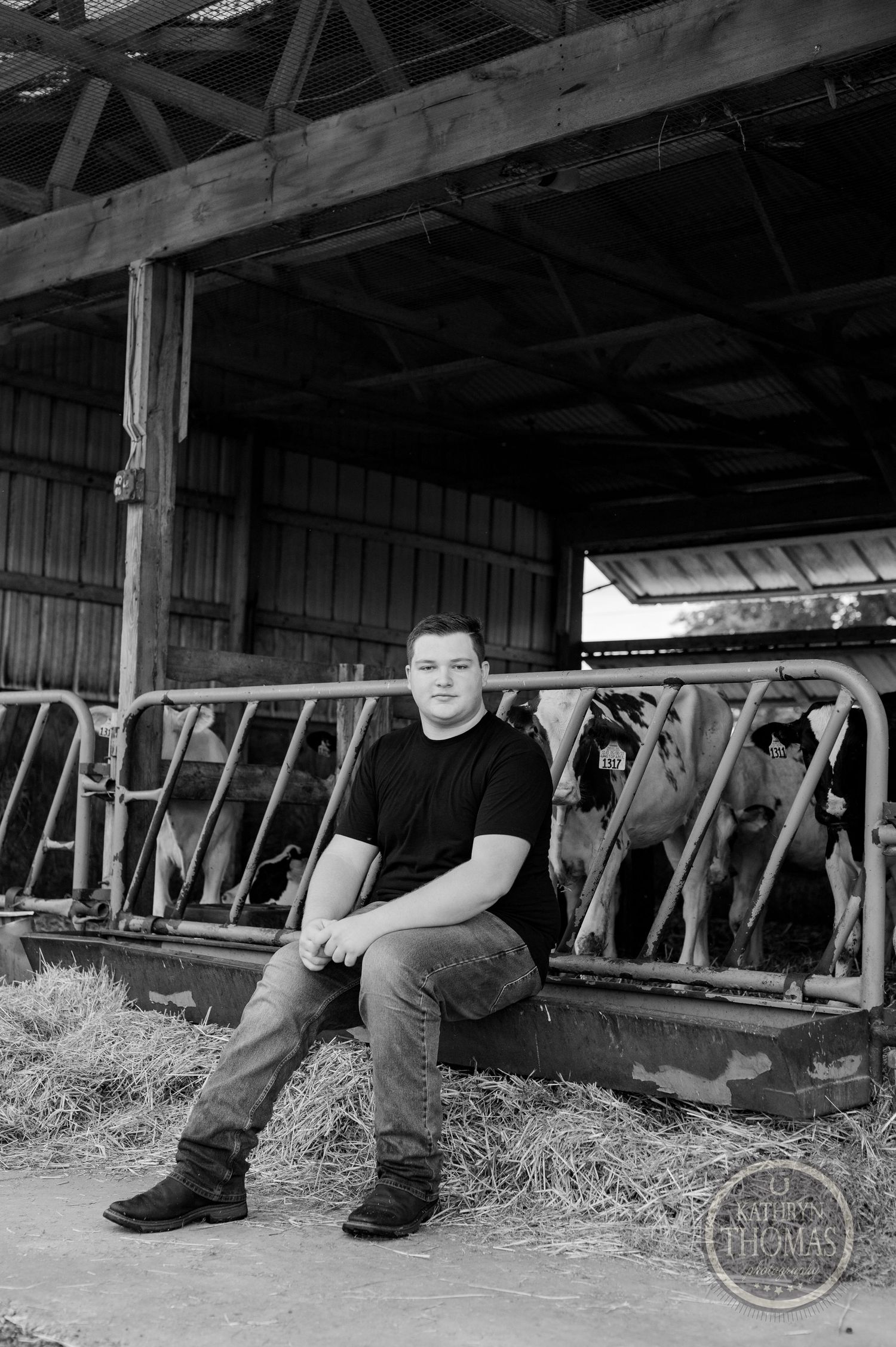 Black and white portrait of someone seated on hay in a dairy barn with cattle stalls visible.