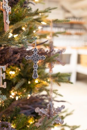 Ornate silver cross ornament hanging on illuminated Christmas tree branches.