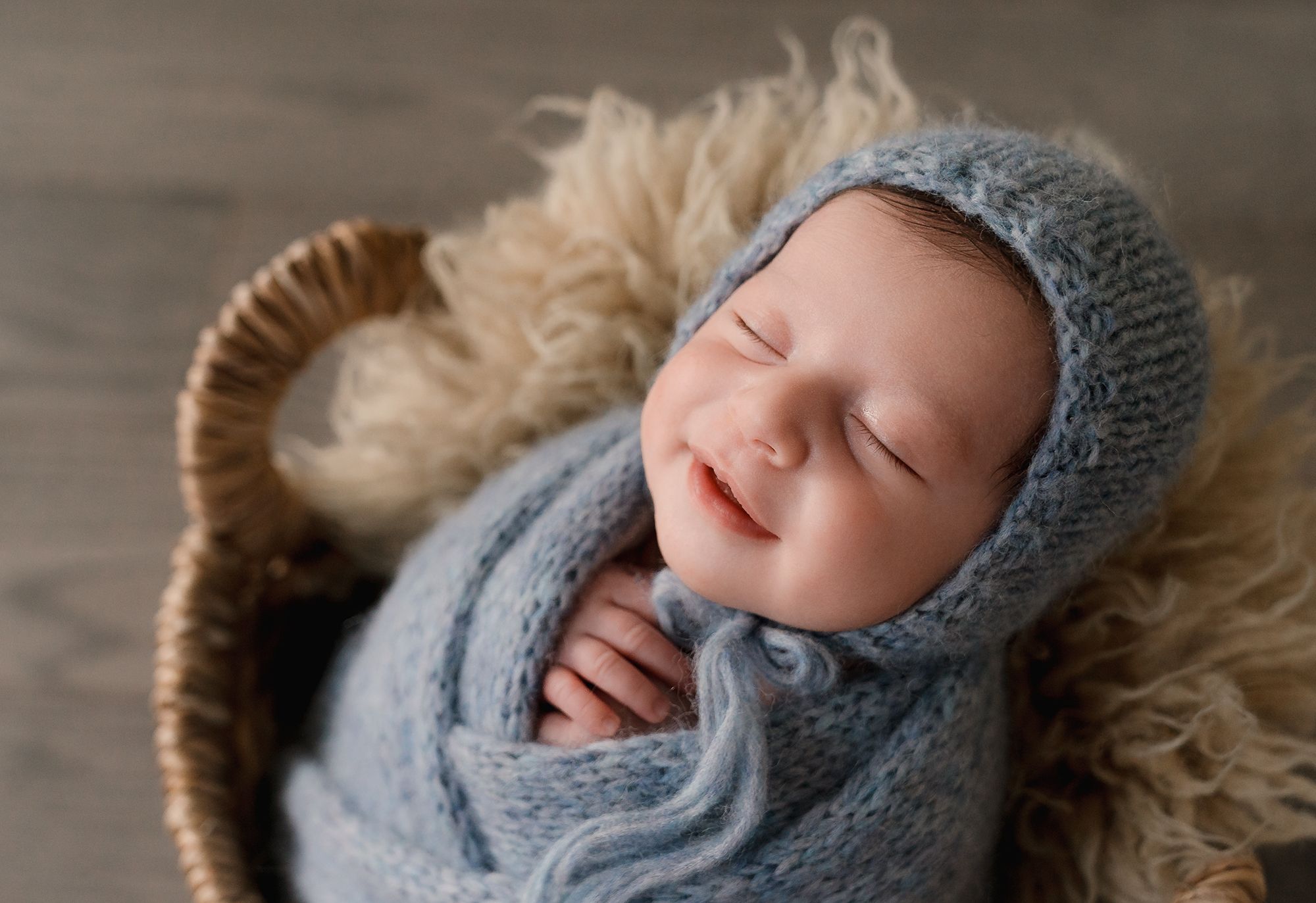 A sleeping newborn baby wrapped in soft gray knit blankets lies on fluffy beige fur.