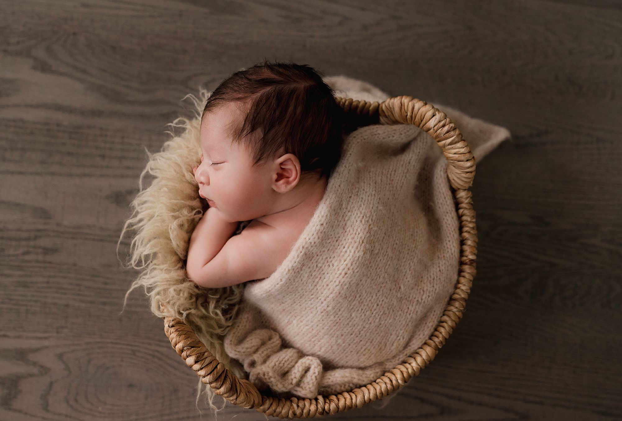 Newborn baby sleeping peacefully in a cozy woven basket with soft beige blanket.
